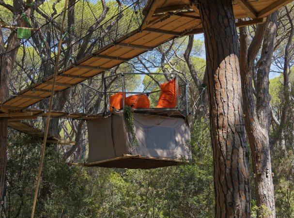 Una tenda sospesa con sedie arancioni sopra, fissata tra gli alberi con passerelle di legno e reti.