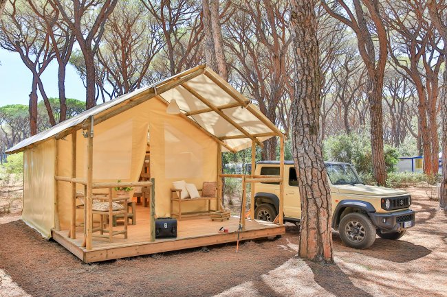Una tenda da campeggio gialla con veranda in legno e un'auto vicina in una pineta.