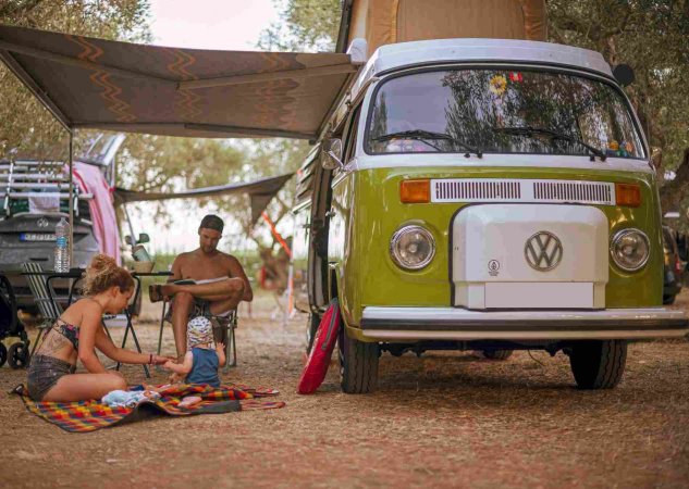 Famiglia in campeggio accanto a un furgone Volkswagen verde, coperta colorata e telo ombreggiante.