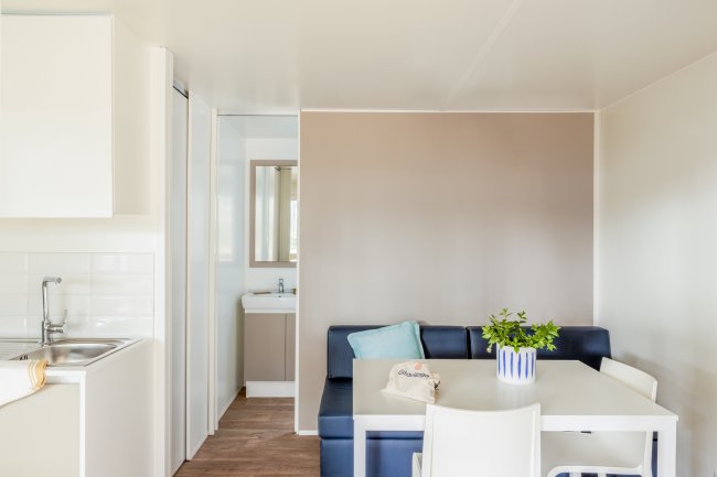 Interno con divano blu, tavolo bianco, piante in vaso, e angolo cottura. Sfondo chiaro.