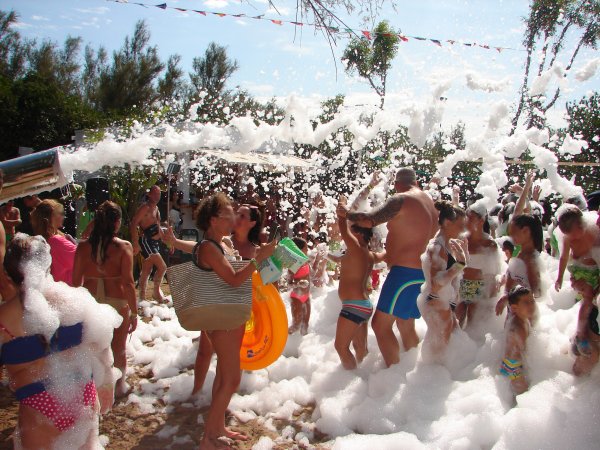 Festa in spiaggia con schiuma, adulti e bambini giocano sorridendo.