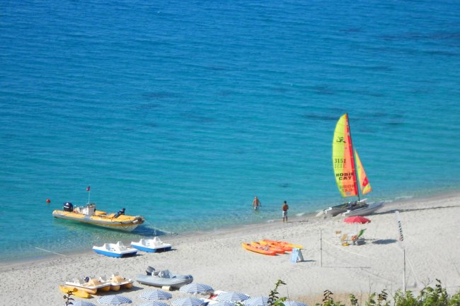Spiaggia con sabbia bianca, barche, pedalò e una barca a vela colorata sul mare turchese.