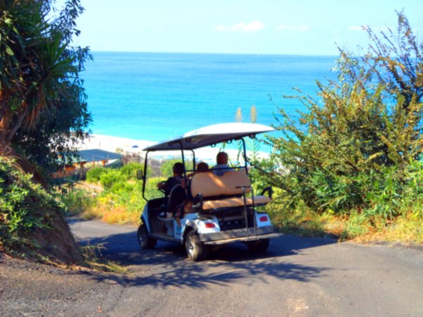 Persone in golf cart su una strada con vegetazione che conduce a una spiaggia e mare blu.