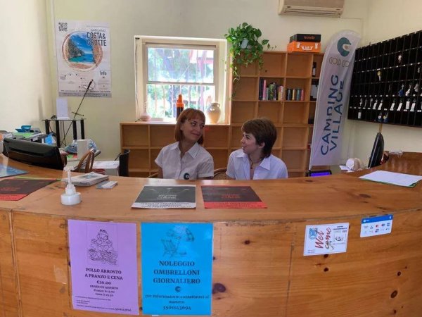 Due persone dietro un banco di accoglienza con volantini e scaffali sullo sfondo.