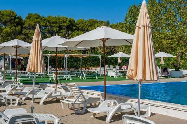 Piscina con lettini bianchi e ombrelloni su un patio, circondata da alberi verdi.