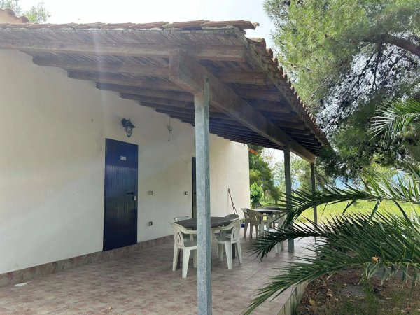 Veranda con tettoia, sedie bianche e tavolo al lato di una casa bianca.