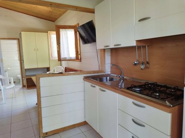 Cucina con mobili bianchi e piano cottura, lavello in acciaio, TV a parete e sedie in plastica.