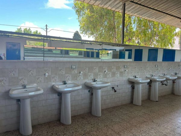 Un bagno all'aperto con file di lavandini bianchi su una parete piastrellata.