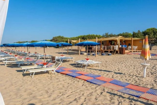 Spiaggia con lettini bianchi, ombrelloni blu e un chiosco in legno sotto un cielo limpido.