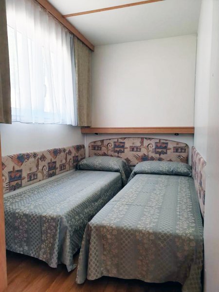 Due letti singoli in una piccola camera con pareti bianche e tende beige.