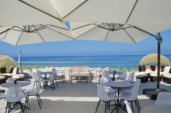 Terrazza con tavoli e sedie sotto grandi ombrelloni, vista sul mare azzurro.