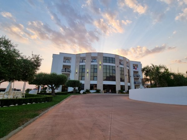 Edificio imponente al tramonto con cielo nuvoloso e luce calda, circondato da alberi e giardini.