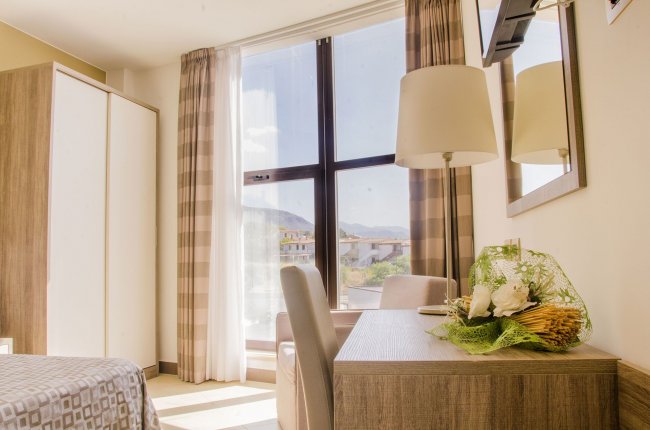Scrivania con fiori, sedia e finestra luminosa con vista esterna. Lampada e TV a parete.