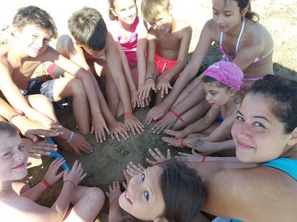 Bambini seduti in cerchio sulla sabbia con le mani a terra, sorridenti.