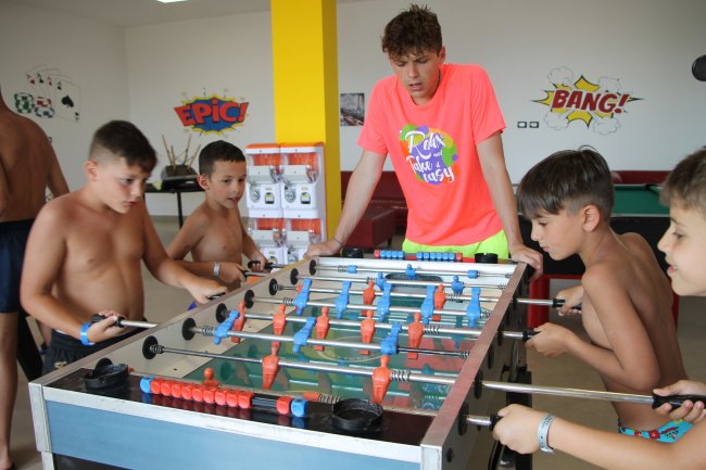 Bambini giocano a biliardino con un adulto in una stanza decorata con scritte colorate.