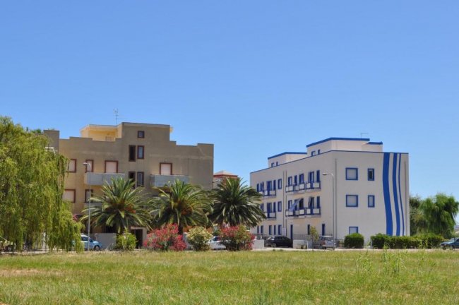 Due edifici, uno bianco-blu e uno più scuro, con palme e spazio verde.