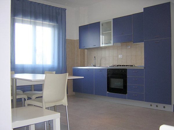 Cucina moderna con mobili blu e tende azzurre, con un tavolo e sedie bianche.
