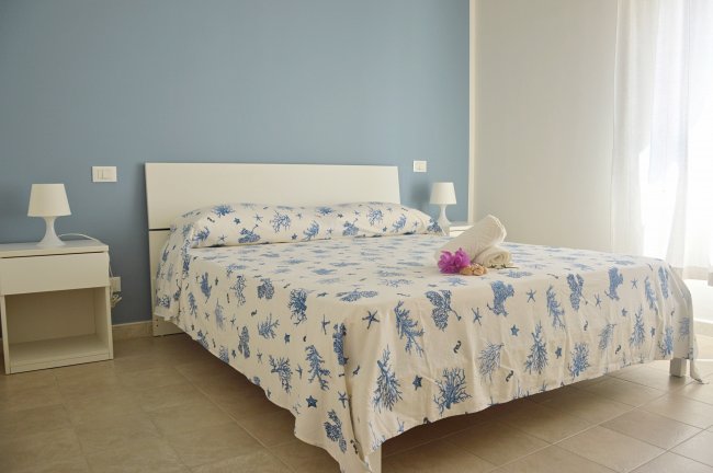 Camera con letto matrimoniale, pareti azzurre, comodini bianchi e lenzuola con stampa marina.