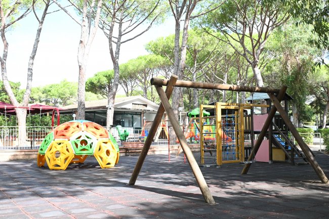 Area giochi con altalena di legno e struttura colorata in parco alberato.