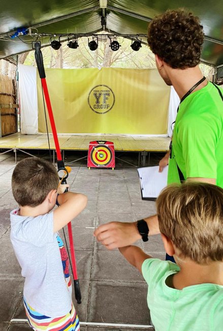 Bambino che pratica tiro con l'arco assistito da un adulto sotto una tenda.