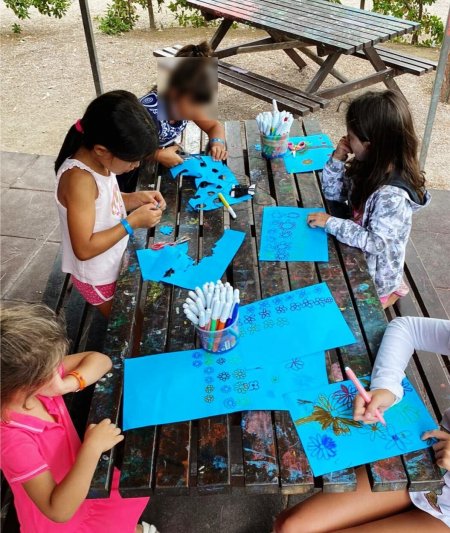 Bambini seduti a un tavolo all'aperto, colorando su fogli blu con pennarelli.
