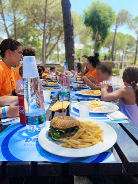 Gruppo di persone a tavola all'aperto; un panino con patatine in primo piano.