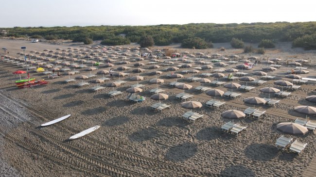 Spiaggia con file ordinate di ombrelloni beige e lettini su sabbia chiara.