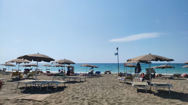 Spiaggia con ombrelloni beige e lettini, mare azzurro in lontananza. Pioggia di mare sullo sfondo.