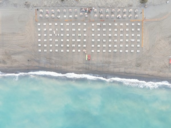 Vista aerea di una spiaggia con file ordinate di ombrelloni e sdraio. Mare verde-azzurro in basso.