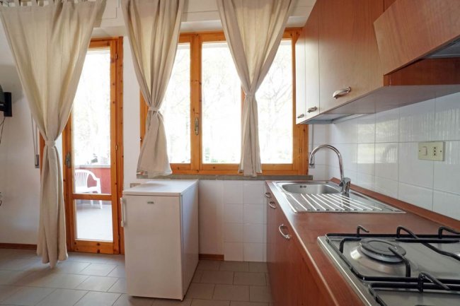 Cucina con lavello, fornelli e mini-frigo, illuminata da una porta e una finestra con tende chiare.