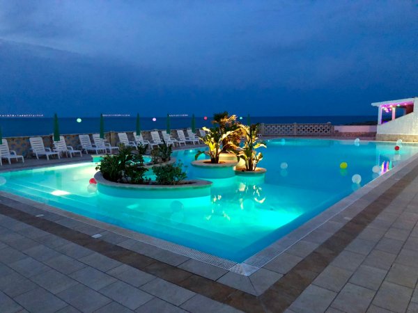 Piscina illuminata, lettini bianchi e mare sullo sfondo.