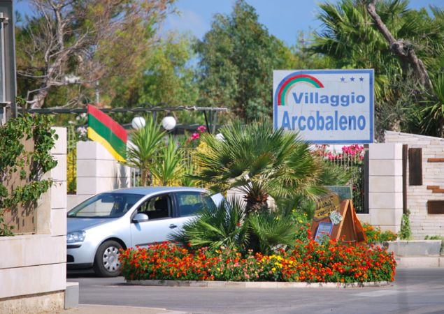 Ingresso di Villaggio Arcobaleno con cartello e auto davanti a un'aiuola fiorita.