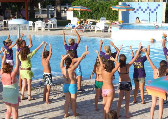 Persone partecipano a una lezione di esercizi accanto a una piscina. Tutti con le braccia sollevate.