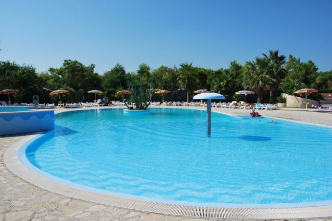 Piscina circolare con ombrelloni e lettini bianchi. Alberi verdi sullo sfondo.