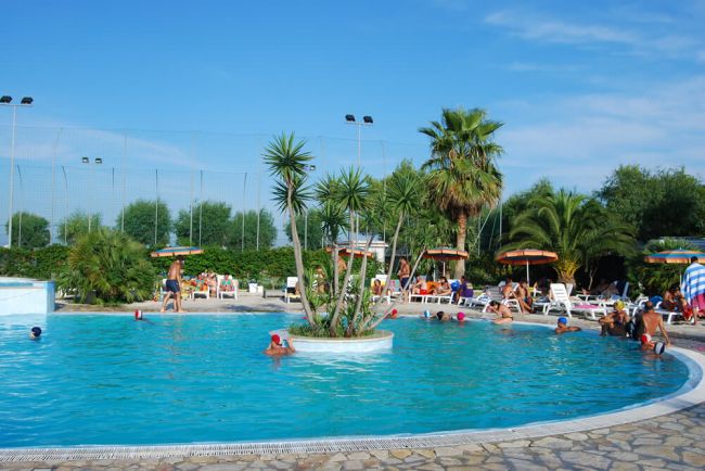 Piscina con centro vegetato, persone nuotano intorno. Palme e ombrelloni sul bordo.