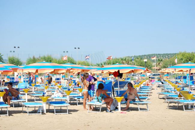 Spiaggia con lettini blu e ombrelloni arancioni, persone rilassate sotto il sole.