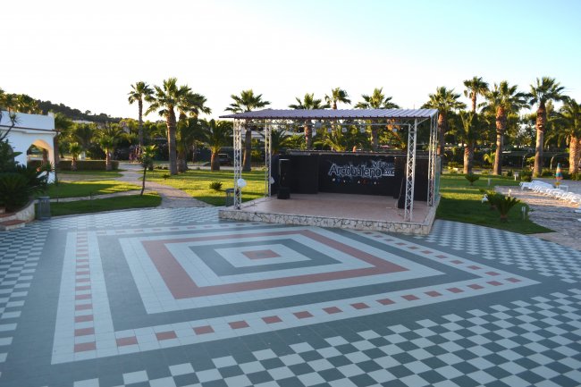 Una piazza con piastrelle a motivi geometrici e un palco tra palme.