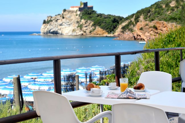 Tavolo bianco con colazione, vista mare e ombrelloni blu su spiaggia.