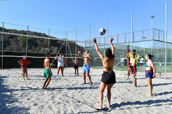 Persone giocano a beach volley su un campo sabbioso recintato, con colline sullo sfondo.