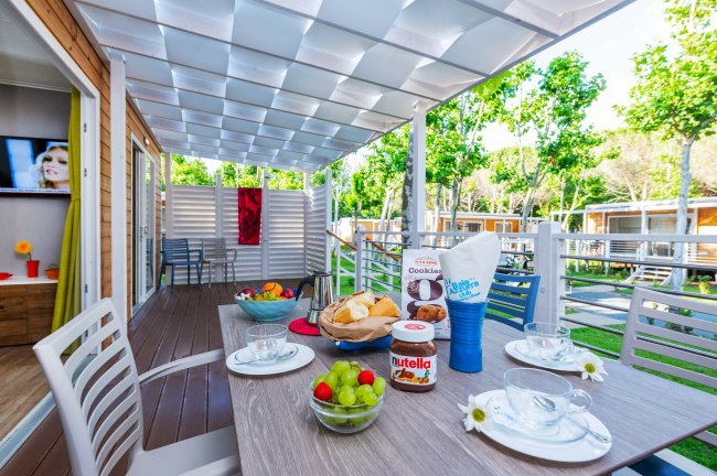 Tavolo all'aperto con colazione preparata, circondato da bungalow e alberi verdi.