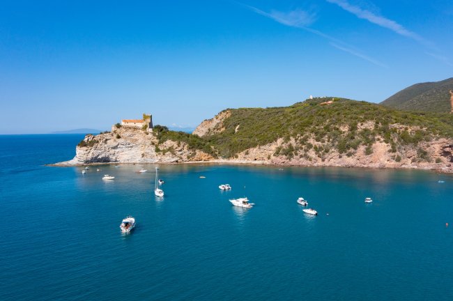 Una baia con barche ormeggiate, scogli e vegetazione, con un edificio su una collina.