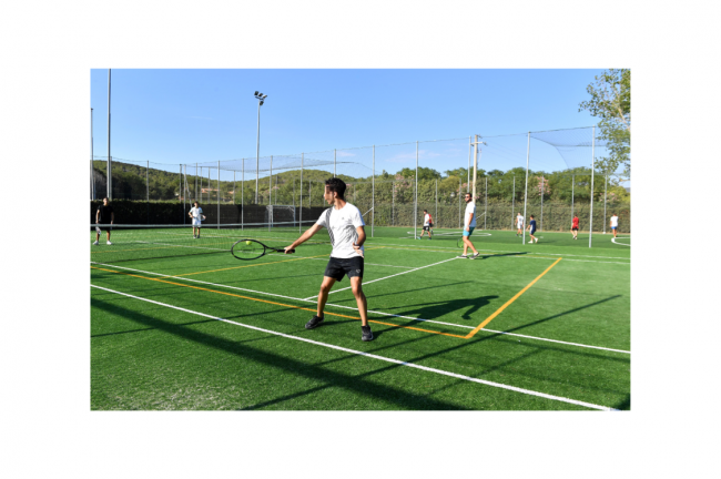 Ragazzo gioca a tennis su un campo all'aperto sotto un cielo limpido. Altri giocatori sullo sfondo.