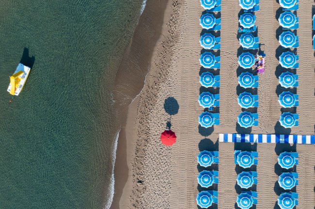 Vista aerea di una spiaggia con ombrelloni blu e un pedalò giallo sull'acqua.