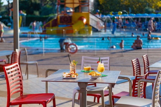 Tavolo con bevande e snack vicino a una piscina affollata.