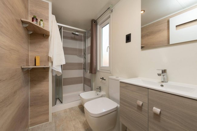 Bagno moderno con doccia in vetro, lavabo, bidet e finestra, rifiniture in legno chiaro.