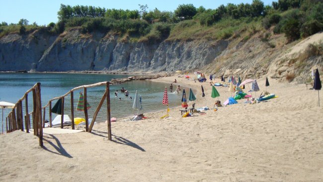 Una spiaggia di sabbia con ombrelloni colorati e persone che nuotano. Scogliere verdi sullo sfondo.