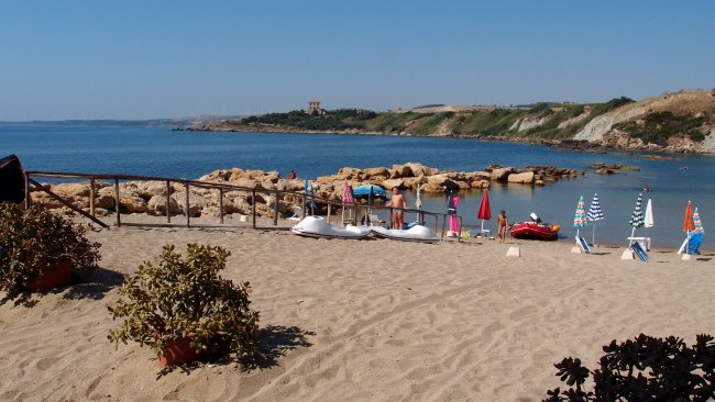 Spiaggia sabbiosa con ombrelloni colorati e pedalò. In lontananza, scogli e colline verdi.