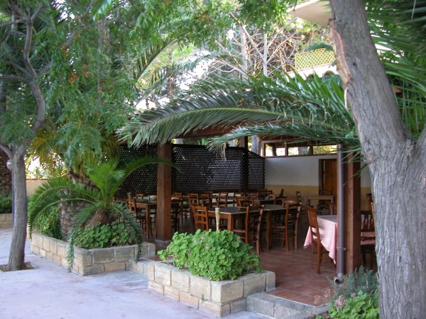 Ristorante all'aperto sotto una veranda con tavoli e sedie in legno, circondato da palme e piante.
