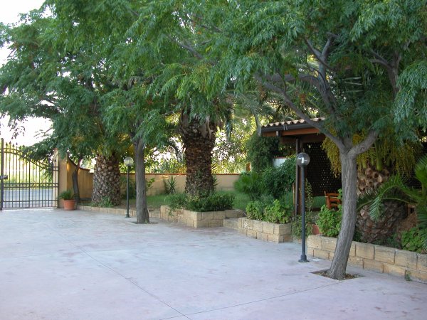 Cortile pavimentato con alberi e aiuole, accanto a un ingresso con cancello.