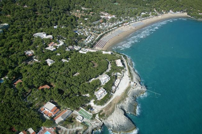 Vista aerea di una costa con spiaggia, mare azzurro e densa vegetazione.